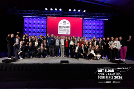 Around 100 people pose for portrait on stage with the text "MIT Sloan Sports Analytics Conference 2026" behind them.
