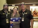 New York firefighters Mike Bellone (left) and Bob Barrett gave an American flag to Gayle Willman (center), who organized trips to Ground Zero for MIT volunteers to care for recovery workers. Behind them are photos taken at the World Trade Center disaster site.