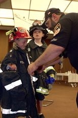 World Trade Center Task Force Fire Safety Director Mike Bellone helps Jamie Joyce, 8, into one of the jackets he and his fellow workers wore in their rescue efforts at the World Trade Center. Jamie's brother Patrick, 10, stands nearby, outfitted in a similar jacket and hat. The children's great-grandfather was a Somerville firefighter. Their great-aunt Kathy Doyle works at Sloan School.