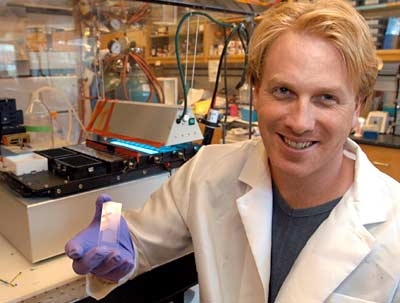 MIT research associate Dan Anderson holds a slide patterned with some 1,700 spots of biomaterial upon which he grows human embryonic stem cells. The machine behind him is the robot key to patterning and analyzing the slide.