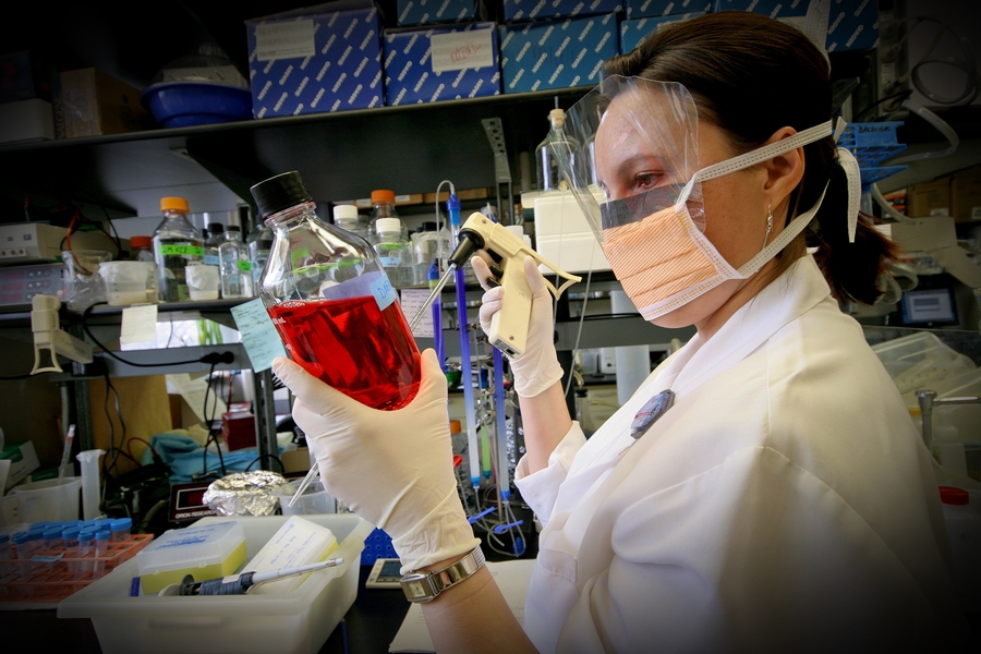 Woman in a lab wearing personal protective equipment and holding a bottle of red liquid