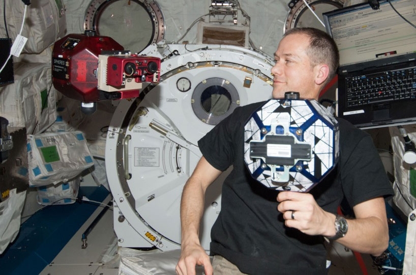 Aboard the International Space Station, astronaut Tom Marshburn works with an MIT Space Systems Lab SPHERES micro satellite. The visually enabled SPHERE is the red object on the left — it's scrutinizing a target SPHERE that Marshburn's holding. 