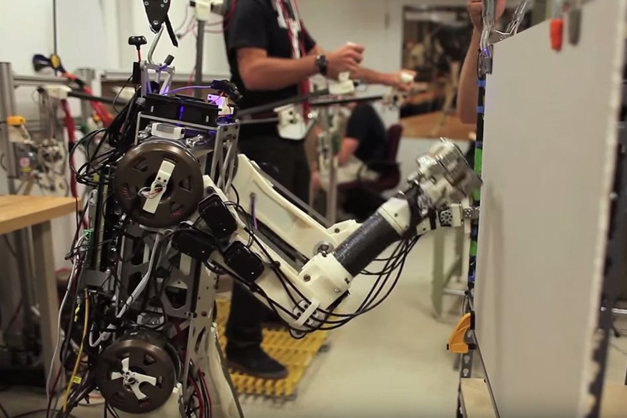 HERMES, a bipedal robot (foreground) designed for disaster response, punches through drywall while keeping its balance, guided by a human operator (background). 