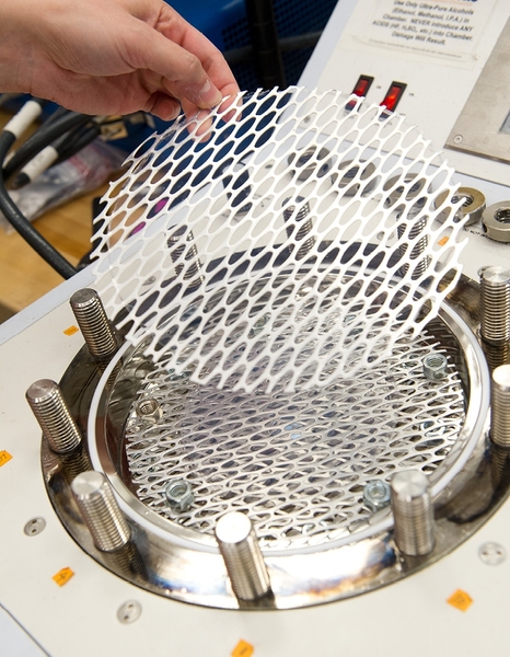 A completed aerogel disk is carefully lifted out of the pressure vessel, its interior pores now filled with air. The disk is almost invisible on top of the metal mesh that supports it. 
