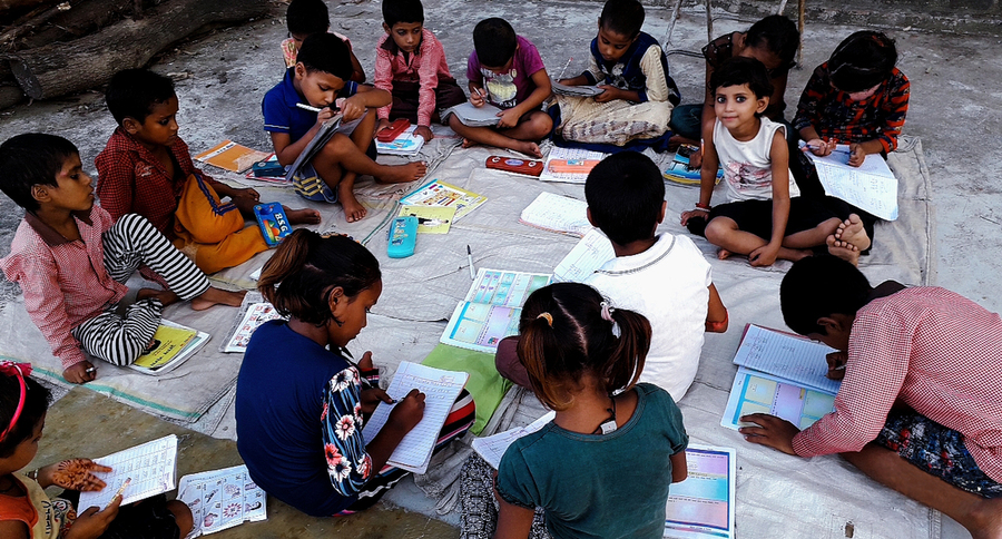Photo of about a dozen young children sitting on the ground doing schoolwork.