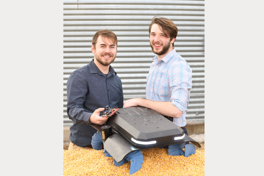 Photo of University of Nebraska Omaha undergraduate students Benjamin Johnson and Zane Zents with their Grain Weevil robot that is sitting on top of a pile of grain.