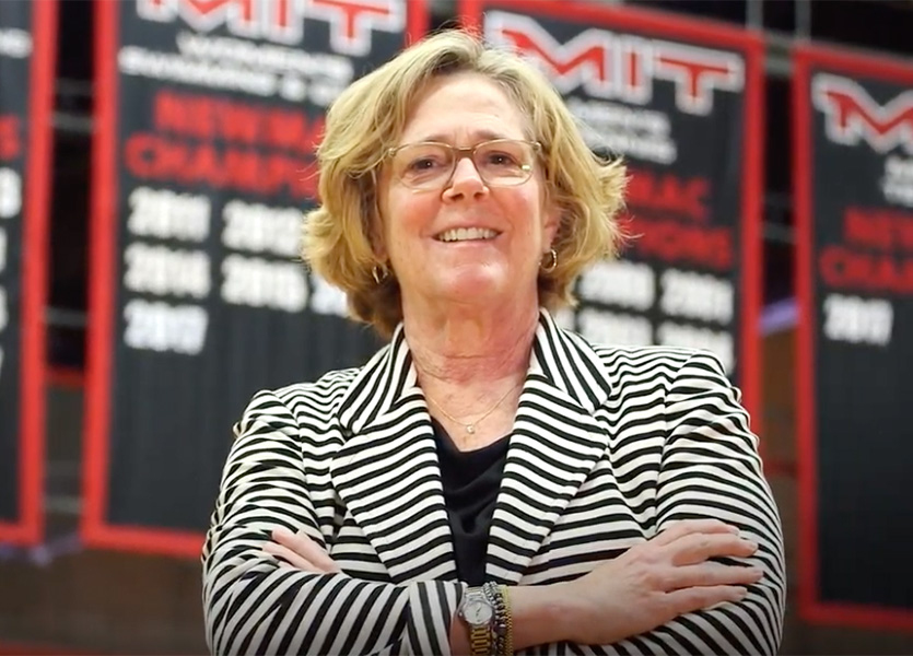 Photo of Julie Soriero standing with her arms folded, in front of championship banners hanging in an MIT gym