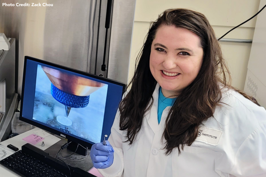 Photo of Nicole Black in the lab, in front of a computer monitor that is displaying a picture of her invention being 3D printed. She also holds her small invention in tweezers.