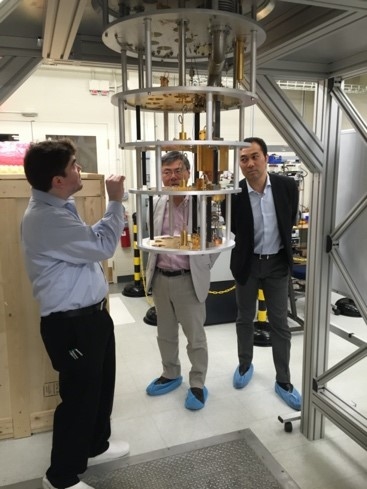 Photo of three researchers in a lab observing a device that looks a bit like an upside-down wedding cake hanging from the ceiling