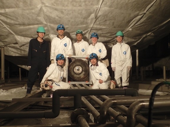 Photo of a group of students and professor in clean suits and hard hats posing with what looks like a fan, with large pipes running along the ground in front of them