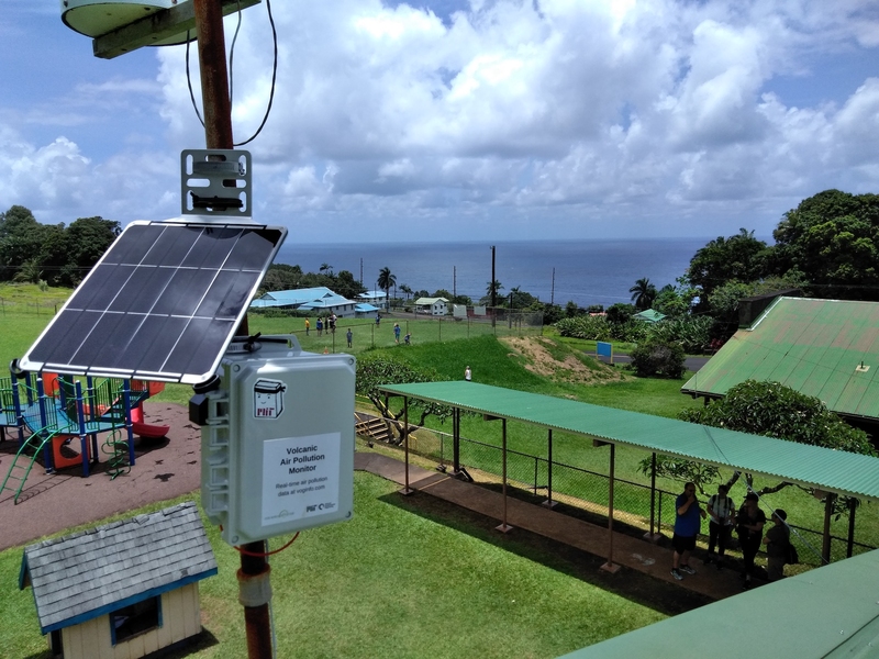 Photo of a solar-powered air quality sensor, labeled with a sticker that says "volcanic air pollution monitor," installed next to a playground and park next to the ocean