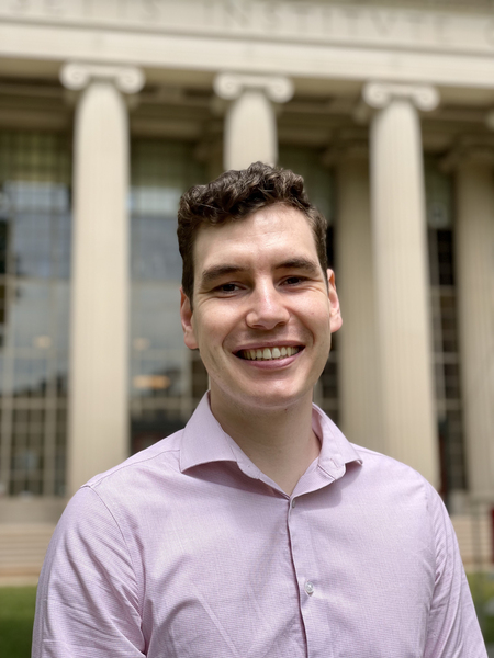 Photo of a man smiling, wearing a purple shirt, and standing in front of columns decorating a large building.