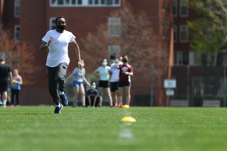 Photo of an MIT student in white shirt and black athletic pants running down Briggs Field in ring toss event.