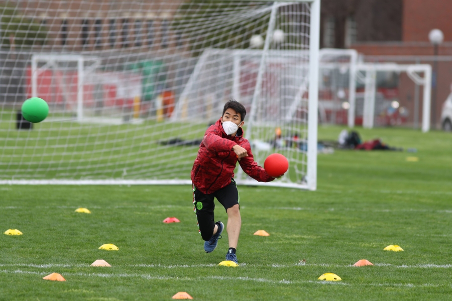 Photo of an MIT student wearing red jacket and black pants throwing a green dodgeball and has a red dodgeball in hand.