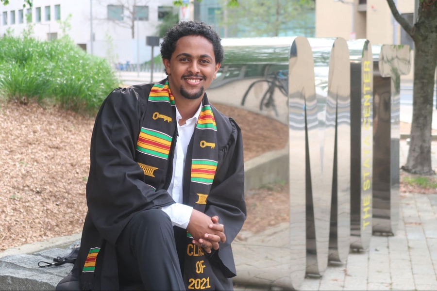 Young man in graduation apparel sits in front of a silver statue featuring the letters M-I-T