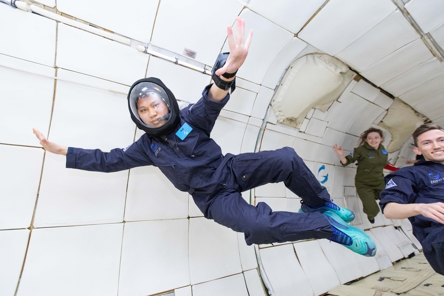 Photo of a woman in a blue jumpsuit and a clear helmet, “flying” in a microgravity environment