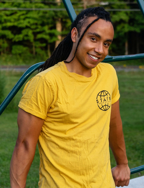 Miles Johnson, with long braids and a bright yellow t-shirt, smiles while leaning on what looks like metal playground equipment in a park