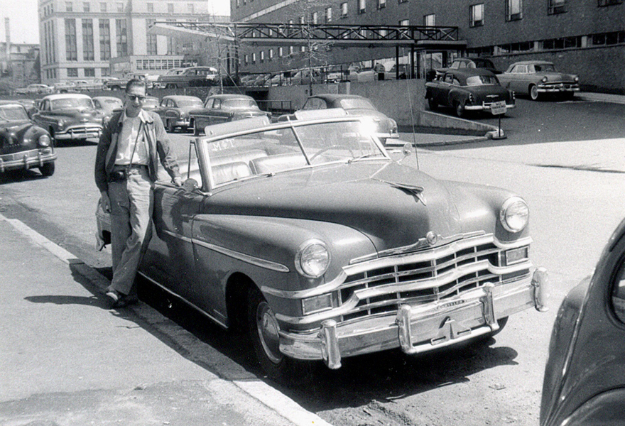 Black and white photo of a young Bob Logcher standing next to a 50s-era car parked on a street, with other similar cars around him