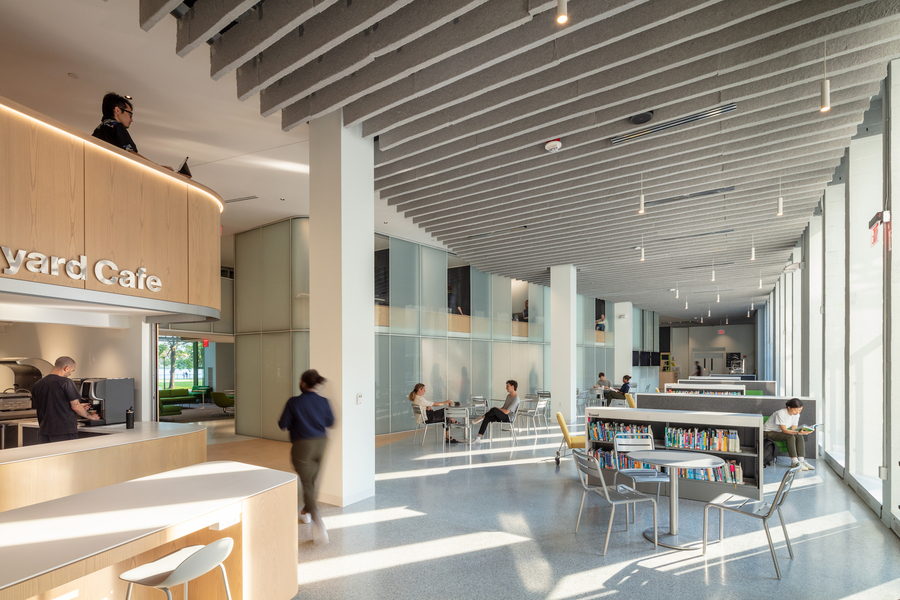 Photo of a person walking by a cafe serving area in the foreground while people are sitting at cafe tables in the background.