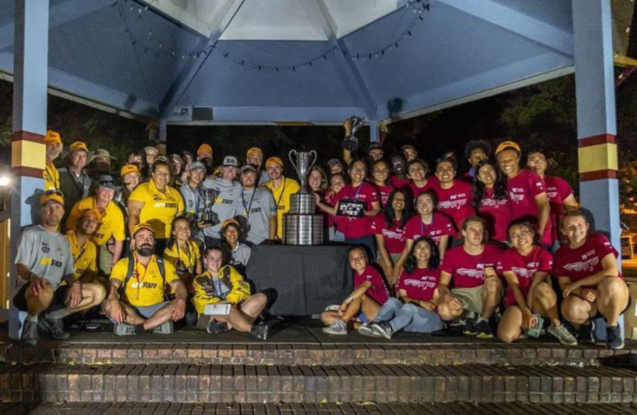 Two teams of college students, one group in yellow t-shirts, one in crimson t-shirts, stand together on a stage, with a large trophy in the middle