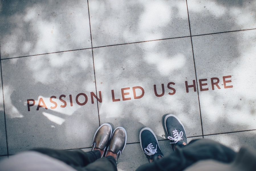 Two pairs of sneakered feet on the sidewalk with the words PASSION LED US HERE