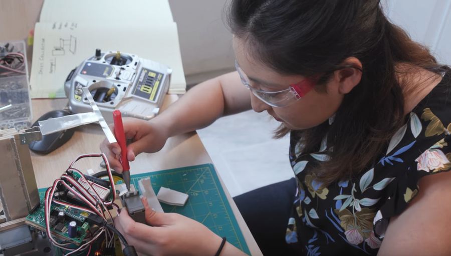Photo of Julianna Rodriguez working on a robot on a kitchen table. She is holding an instrument that looks like a pen, manipulating a small piece of equipment with colorful wires popping out of it.