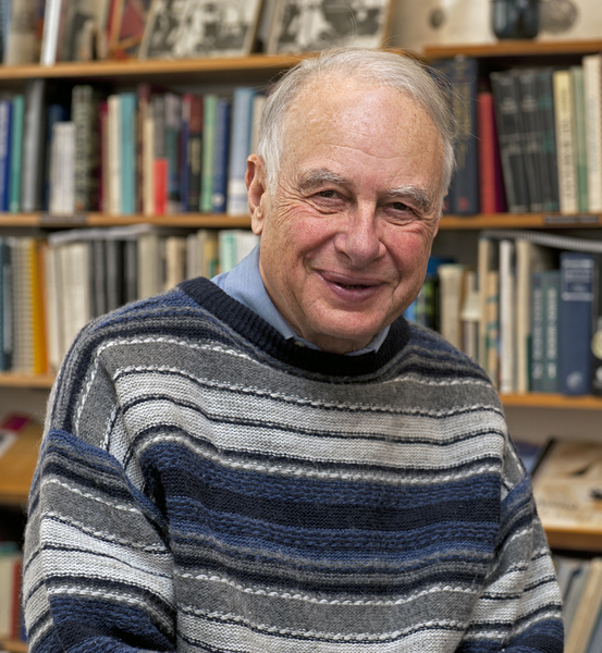 Portrait photo of a smiling man wearing a striped sweater and standing in front of book shelves