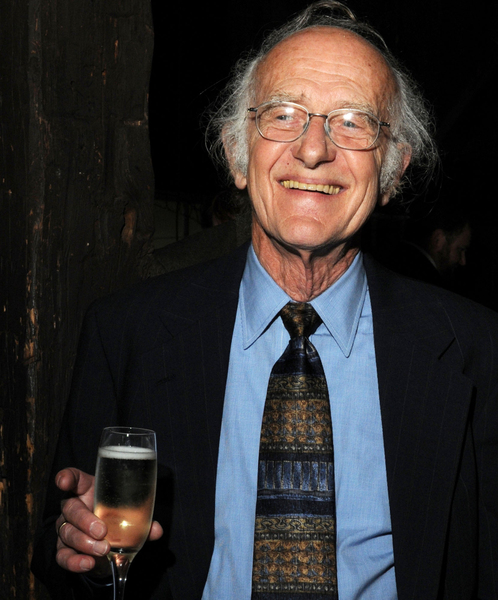 A well-dressed man with silver hair holds a glass of white wine while smiling and looking to his right