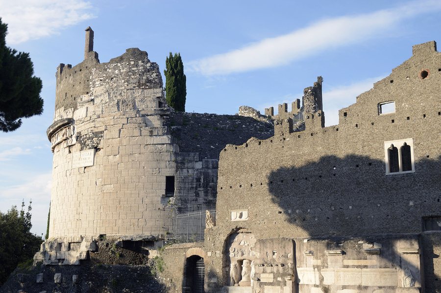 Photo of castle ruins. At left is a circular stone tower partially destroyed and missing a roof. At right is a partially intact facade of brick, also missing a roof, with columns and ancient sculptures embedded toward the bottom.