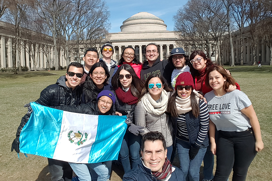 Photo of 14 young people posing on a large field (Killian Court) in front of MIT Building 10, the Great Dome, in winter. One person holds a Guatemalan flag, which features two bars of aqua and a central bar of white with laurels and a bird