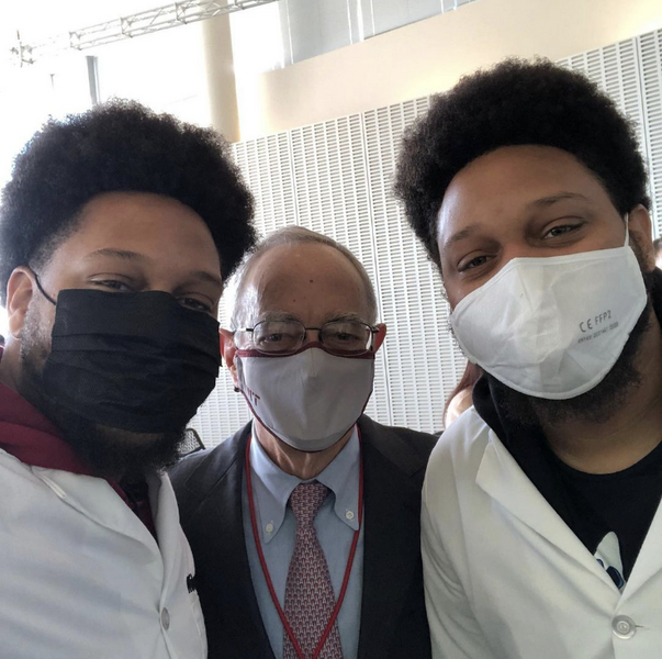 Three men, all wearing face masks, pose for a selfie in an indoor setting with a white wall behind them