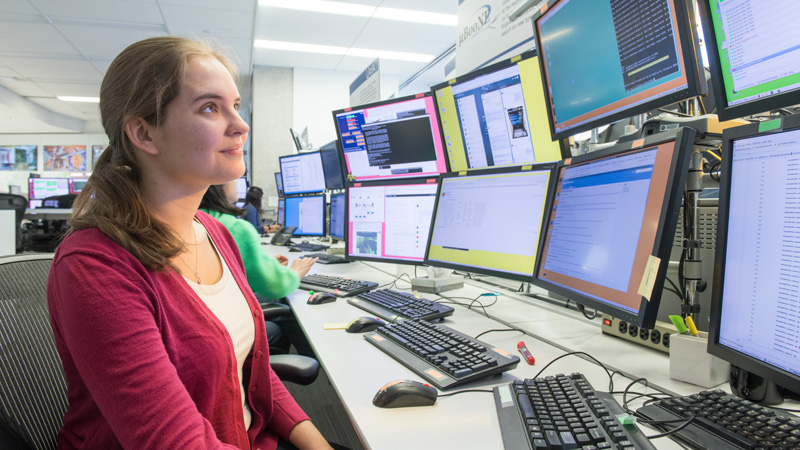 Photo of a young woman seated in a lab with about 10 computer monitors and keyboards showing technical data