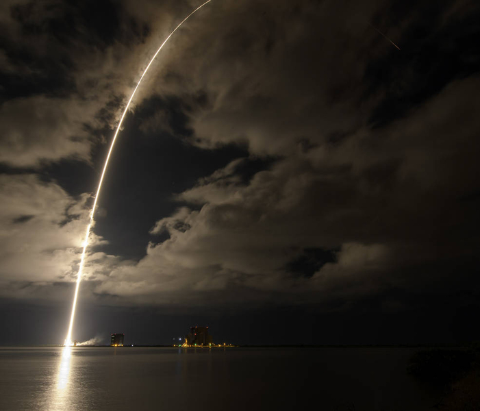 Photo of a bright arc of light reachihng through clouds above a body of water at night
