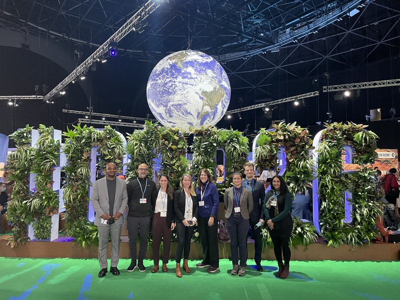 Photo of 8 MIT affiliates stand in a large convention center, in front of a sign made with leaves that reads “#COP26.” A large Earth globe hangs down behind them.