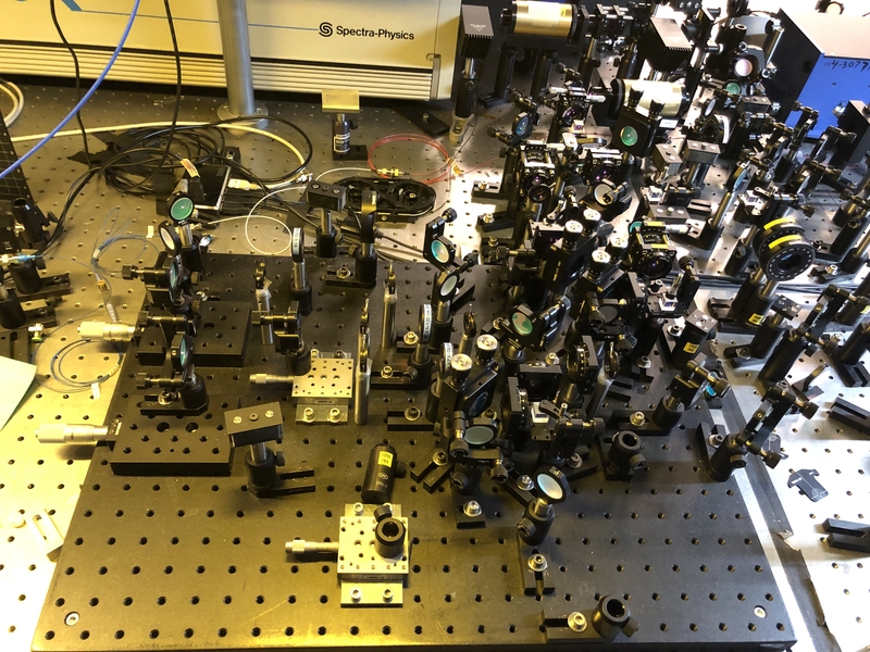 A tabletop showing an optical setup, consisting of more than a dozen mirrors and other hardware elements.