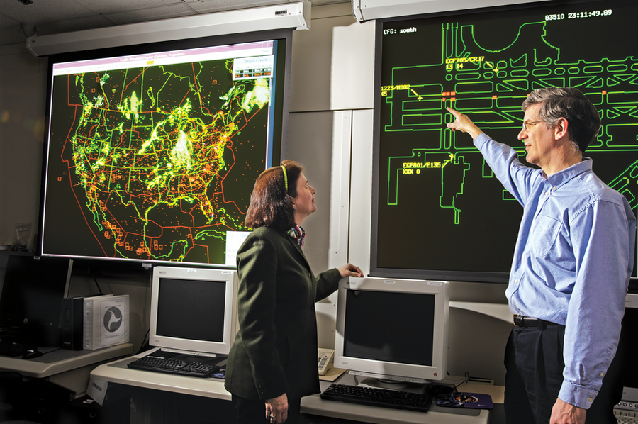 Photo of two researchers standing in a lab with computers in front of two large screens displaying a U.S. map of air traffic and of an airport runway layout.