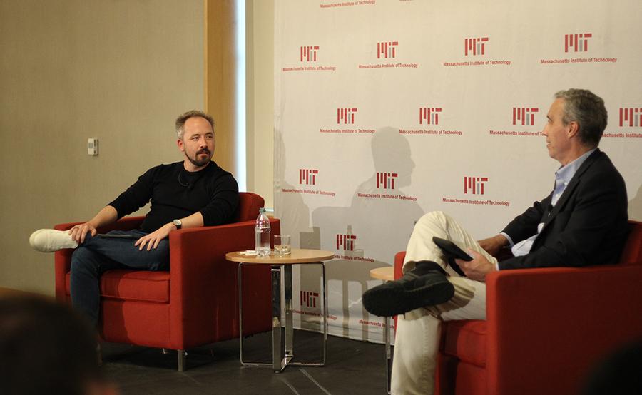 Photo of Drew Houston and Dan Huttenlocher, seated in red chairs and speaking to each other in front of a wall with a repeating MIT logo