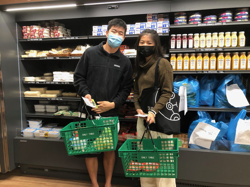Photo of two students in Daily Table, masked and smiling, posing with a basket of grocery items.