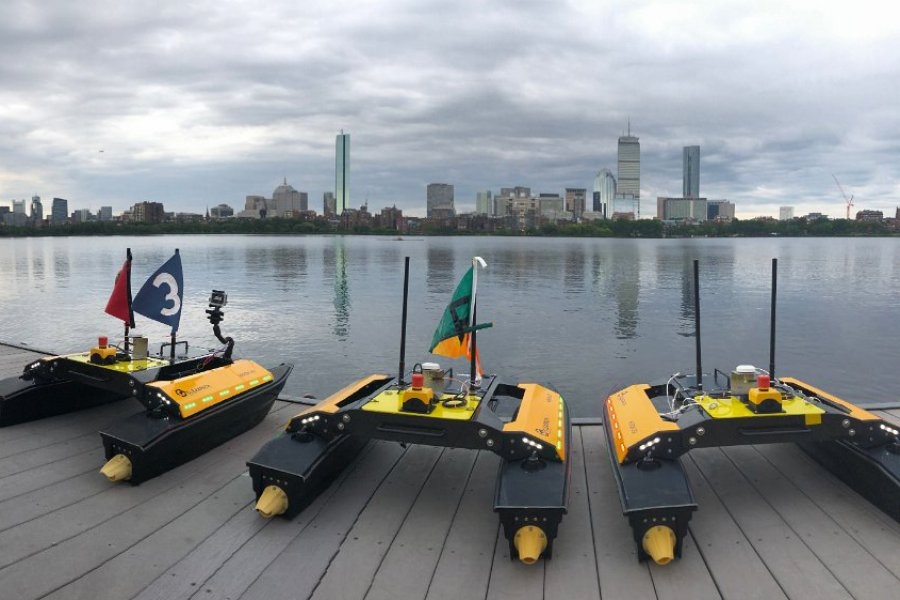 Photo of three autonomous boats parked by the Charles River, with Boston’s skyline in the background