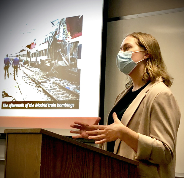 Photo of Nina Miller standing at a podium, wearing a mask and gesturing with her hands
