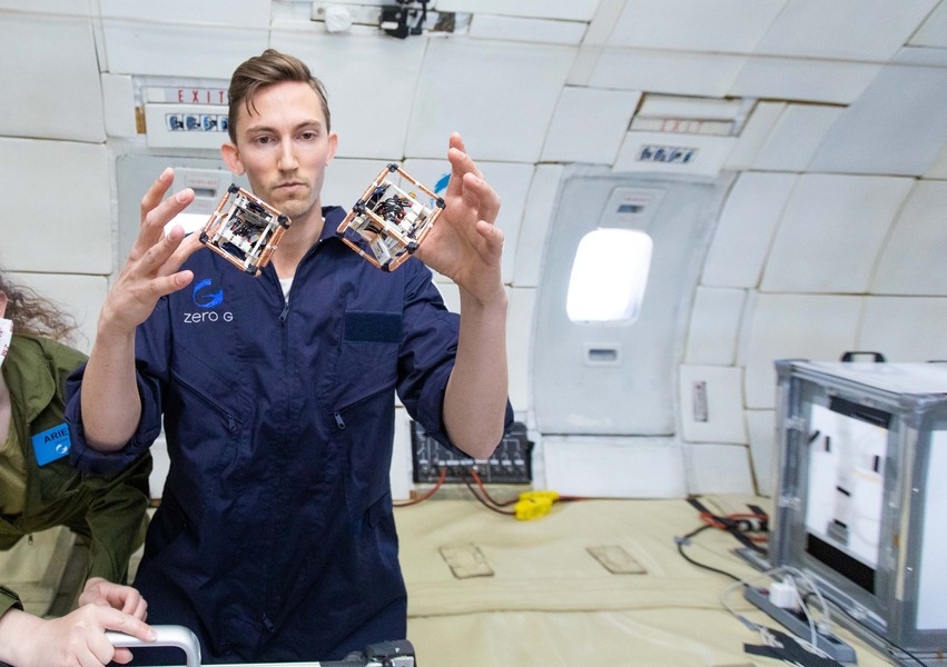 Photo of Martin Nisser in a blue jumpsuit inside a research airplane. Two cubes of electronics float between his hands.