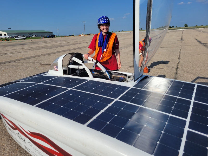 Photo of Cameron Kodesh standing behind solar car beneath a blue sky