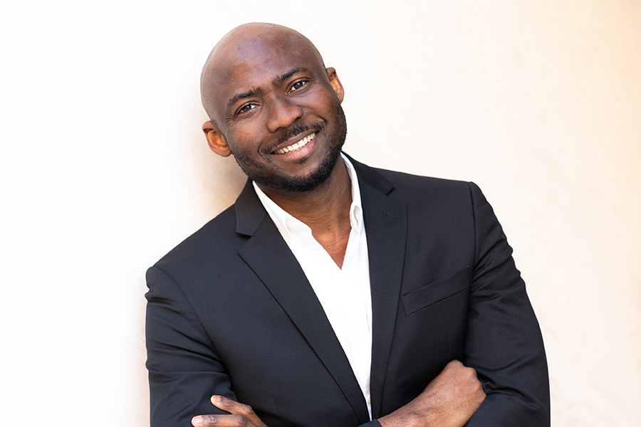 Lawrence Udeigwe stands with arms crossed, standing next to a white wall