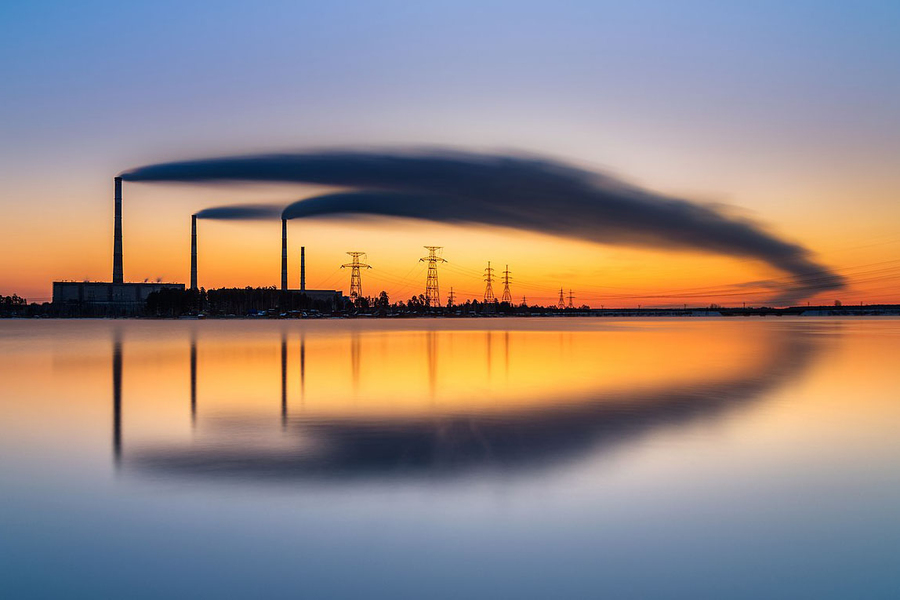 three smokestacks billow symmetric plumes of smoke over the horizon, which is reflected in a pool of still water below.