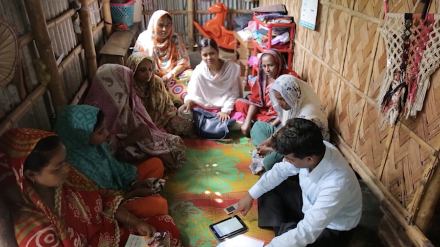 In Bangladesh, a group of eight women sit in a circle in a small room. A man, also sitting on the floor with them, is explaining a BRAC humanitarian program. An iPad and cell phone are on the floor. 