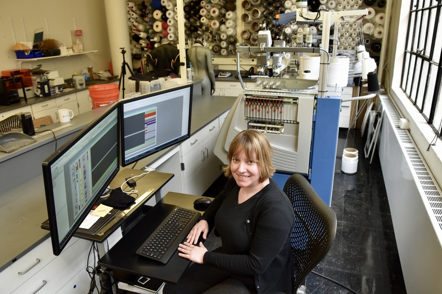 Photo of Lavender Tessmer seated next to a computer with dual screens, and in front of a large knitting machine. On the wall behind her are dozens of spools of thread, mostly black and white but a few in bright colors. Two mannequins also stand in the back of the room.