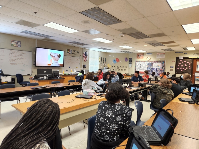 Seventeen middle school students sit in their classroom looking at a TV displaying video lesson about AI