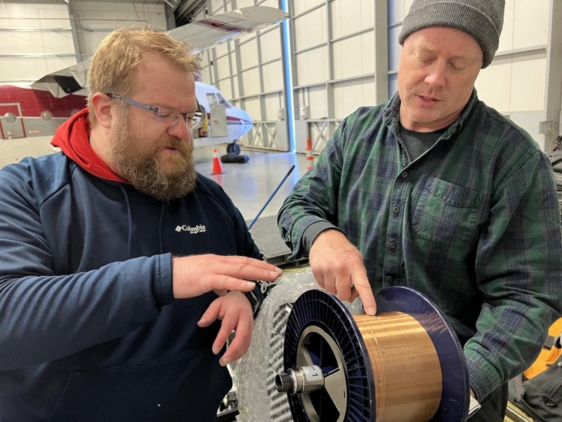 Ben Evans and Dave Whelihan point to and discuss a large spool of copper wire. They are standing inside a hangar with a small airplane behind them. 