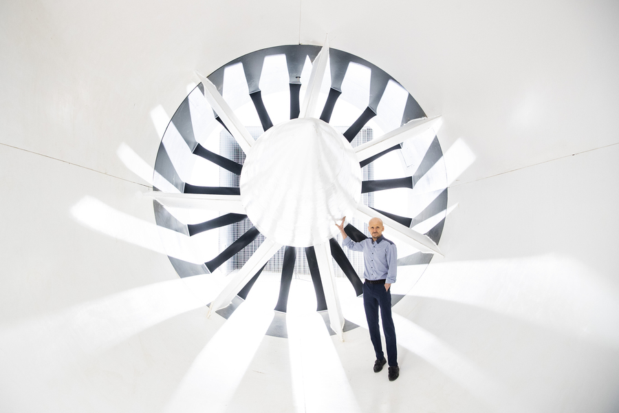 Photo of Mark Drela standing before a large fan inside a white wind tunnel. The scene is backlit with rays of light radiating dramatically toward the camera.