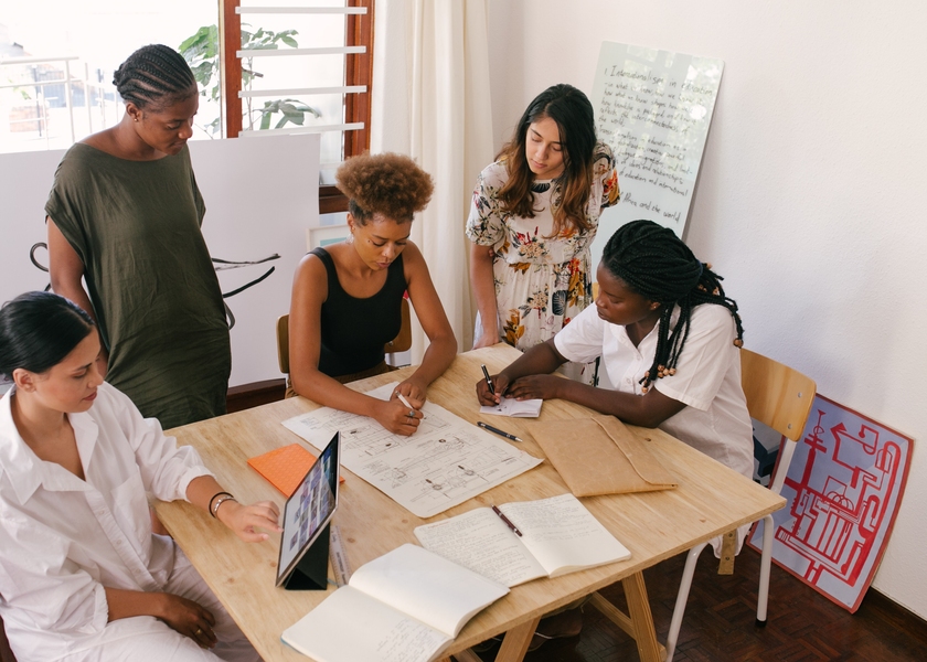 Photo of five women surrounding a table during a meeting. One works on a tablet, two write on paper, and the last two look at their colleagues' work.
